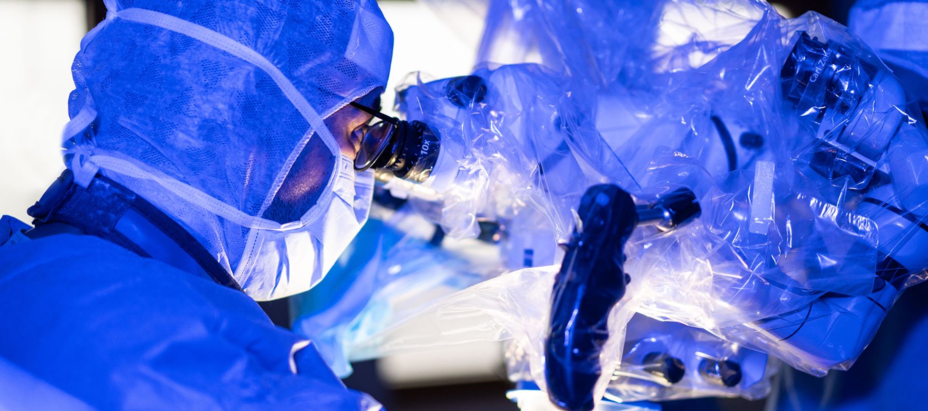 Hr. Alsingaby ist in steriler OP Bekleidung mit Haube und Maske im OP. Er blickt durch ein Mikroskop welches auch mit steriler Folie abgeklebt ist. Die Umgebung ist blau beleuchtet.