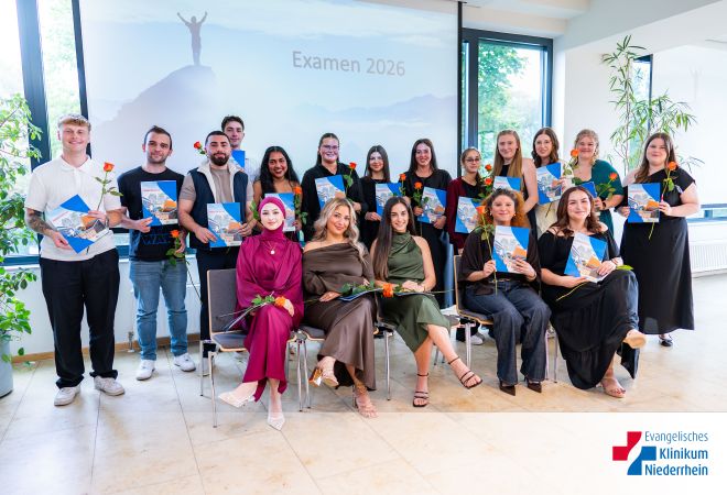 18 Absolventinnen und Absolventen der Pflegeschule, eine Reihe sitzend, dahinter weitere stehend; alle halten Zeugnis-Mappe und Rose, im Hintergrund Leinwand mit ‚Examen 2026‘.