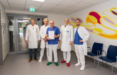 Gruppenbild auf dem Stationsflur. 5 Ärzte stehen nebeneinander, einer von ihnen hält ein Zertifikat in die Kamera. Alle lächeln. Im Hintergrund ist eine gelb-rote Wandmalerei zu sehen.