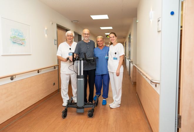 Vier Personen auf dem Stationsflur des Evangelischen Klinikums Niederrhein: Eine Ärztin, ein Arzt und eine Pflegekraft stehen lächelnd neben einem Patienten, der sich an einem modernen Gehwagen stützt. Alle lächeln. Der Flur ist hell und freundlich.