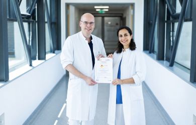 Ein Arzt und eine Ärztin präsentieren in einem Verbindungsgang des Krankenhauses das Zertifikat zur Auszeichnung des Evangelischen Klinikums Niederrhein als Cardiac Arrest Center.