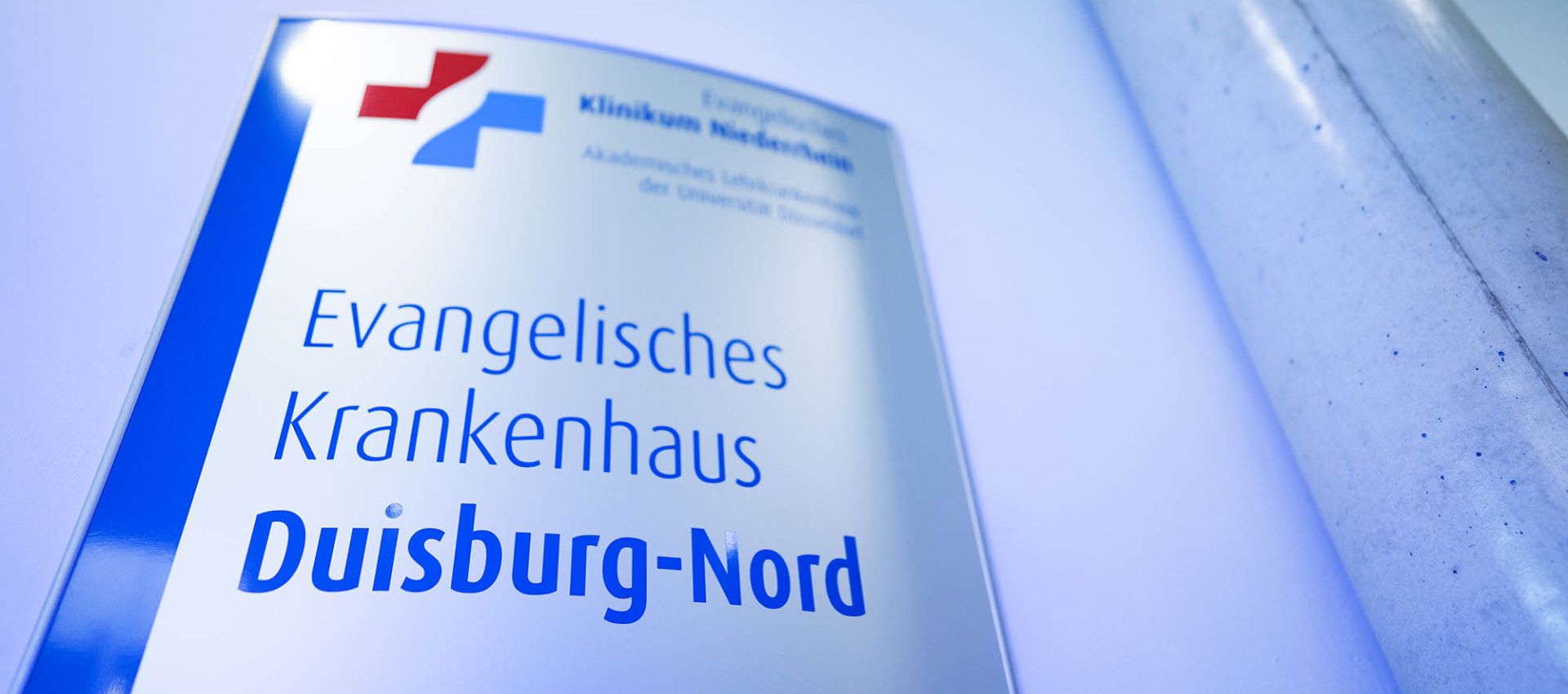 Ein Schild im Eingangsbereich zeigt den Schriftzug „Evangelisches Krankenhaus Duisburg-Nord“ in blauer Unternehmensfarbe. Dadrüber ist das rot-blaue Kreuz als Logo des Klinikverbundes zu sehen.