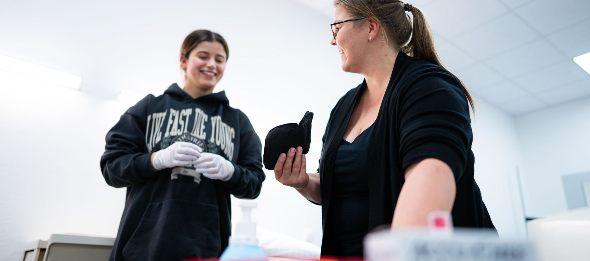 Eine Ausbilderin zeigt einer Auszubildenden etwas auf einem Tablet. Beide lächeln. Die Auszubildende trägt Einweghandschuhe, im Vordergrund sind Pflegeutensilien unscharf zu sehen. Die Szene zeigt eine positive Lernatmosphäre im Praxisunterricht.