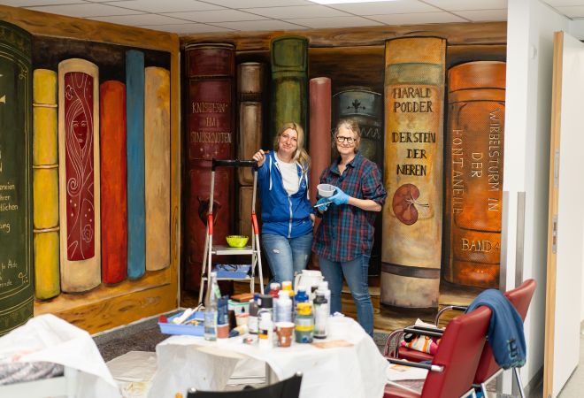 Zwei Frauen stehen mit Malutensilien vor einer großflächigen Wandmalerei mit überdimensionalen Bücher-Motiven im FKlinikflur. Vor ihnen stehen Farbeimer und Pinsel und daneben eine Leiter. Sie tragen Alltagskleidung.