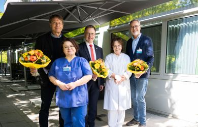 Fünf Personen stehen auf der Terrasse der Palliativstation. Drei halten Blumensträuße in den Händen, sie tragen Alltags- und Businesskleidung. Zwei Mitarbeiterinnen tragen Pflegekleidung. Im Hintergrund sind Sonnenschirme zu sehen.