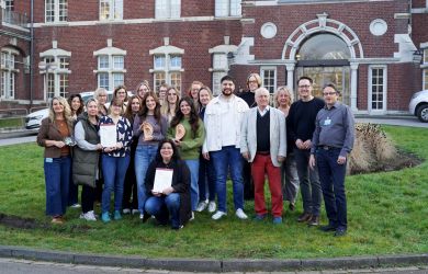 Eine Gruppe von rund zwanzig Teilnehmenden der Akupunktur-Weiterbildung steht gemeinsam auf einer Wiese vor dem historischen Gebäude des Johanniter-Krankenhauses Oberhausen. Einige halten Zertifikate oder Anschauungsmodelle eines Ohrs hoch und lächeln.