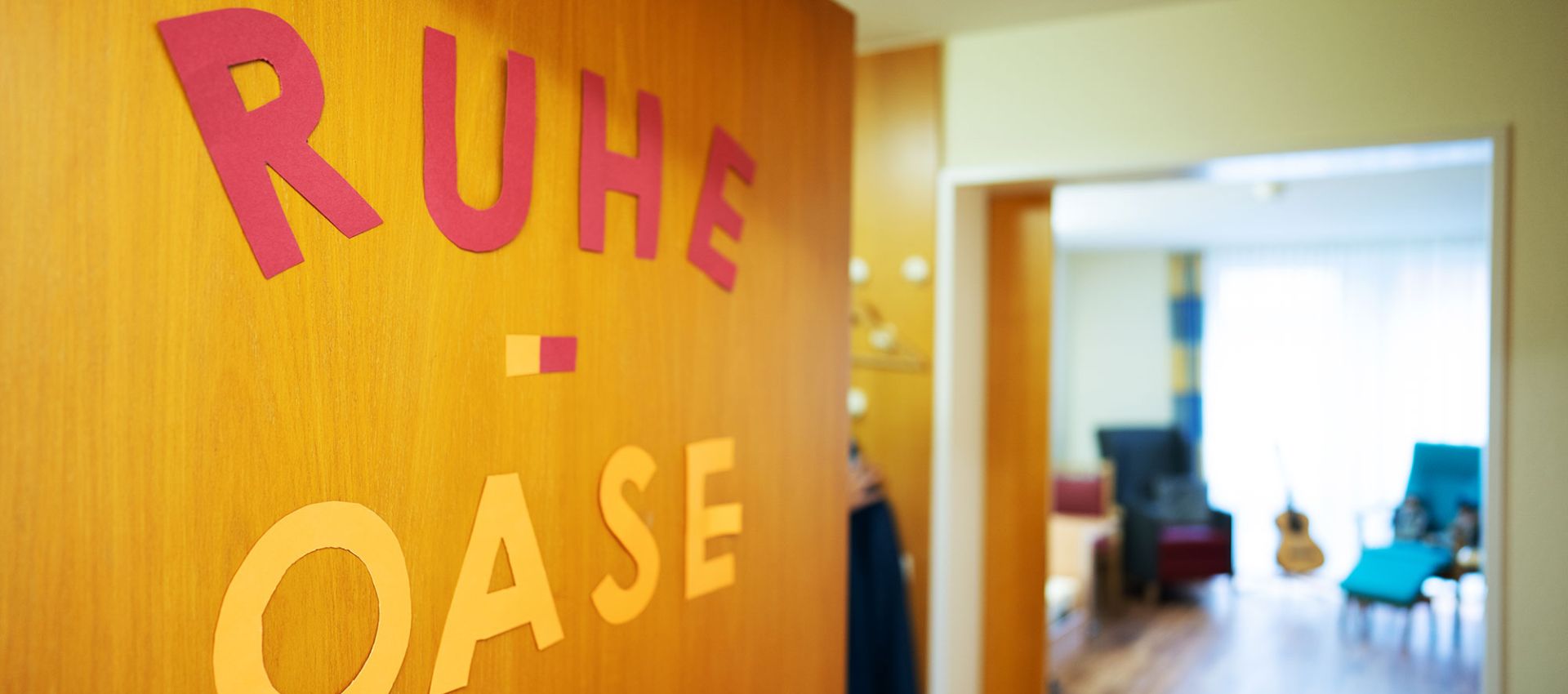 Im Vordergrund eine geöffnete Tür mit dem Wort „Ruheoase“ aus rot-gelben Buchstaben. Dahinter ein einladender, heller Raum mit gemütlichen Sitzgelegenheiten und großen Fenstern..