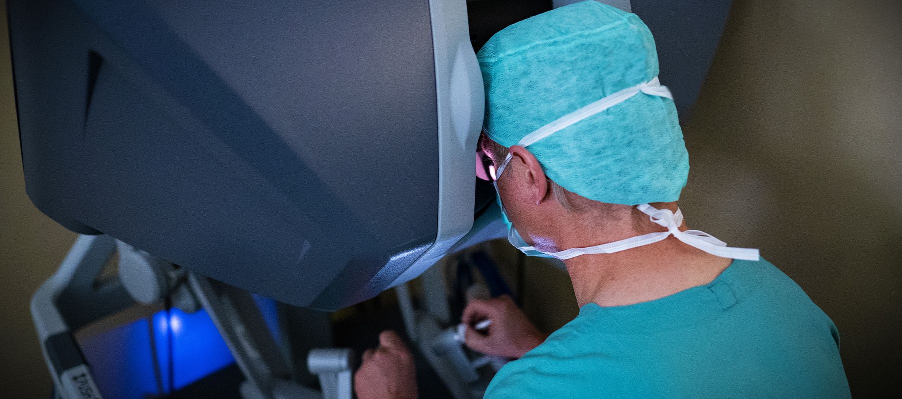 In einem sterilen Operationssaal sitzt Professor Börgermann konzentriert an einer OP-Roboter-Steuerkonsole. Er trägt grüne OP-Kleidung, einschließlich Haube und Mundschutz.