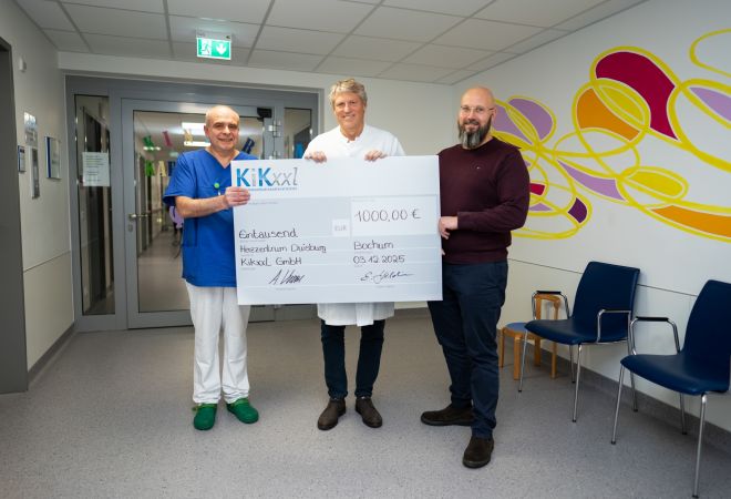 Dr. Gleb Tarusinov aus der Kinderkardiologie, Dr. Michael Scheid (Kinderherzchirurgie) und Marcel Radomski (KiKxxL) stehen im Krankenhausflur und halten gemeinsam einen großen symbolischen Spendencheck über 1000 Euro.