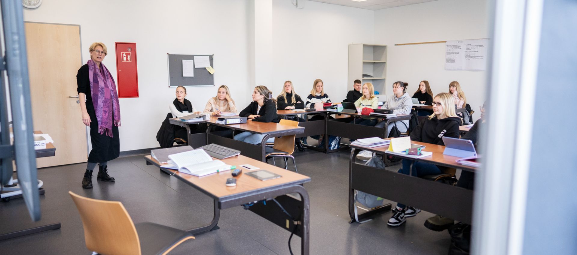 Im Unterrichtsraum der Pflegeschule spricht eine Lehrkraft vor einer Gruppe Auszubildender, die an Tischen sitzen. Die Lernenden sind aufmerksam, der Raum ist hell und klar strukturiert.