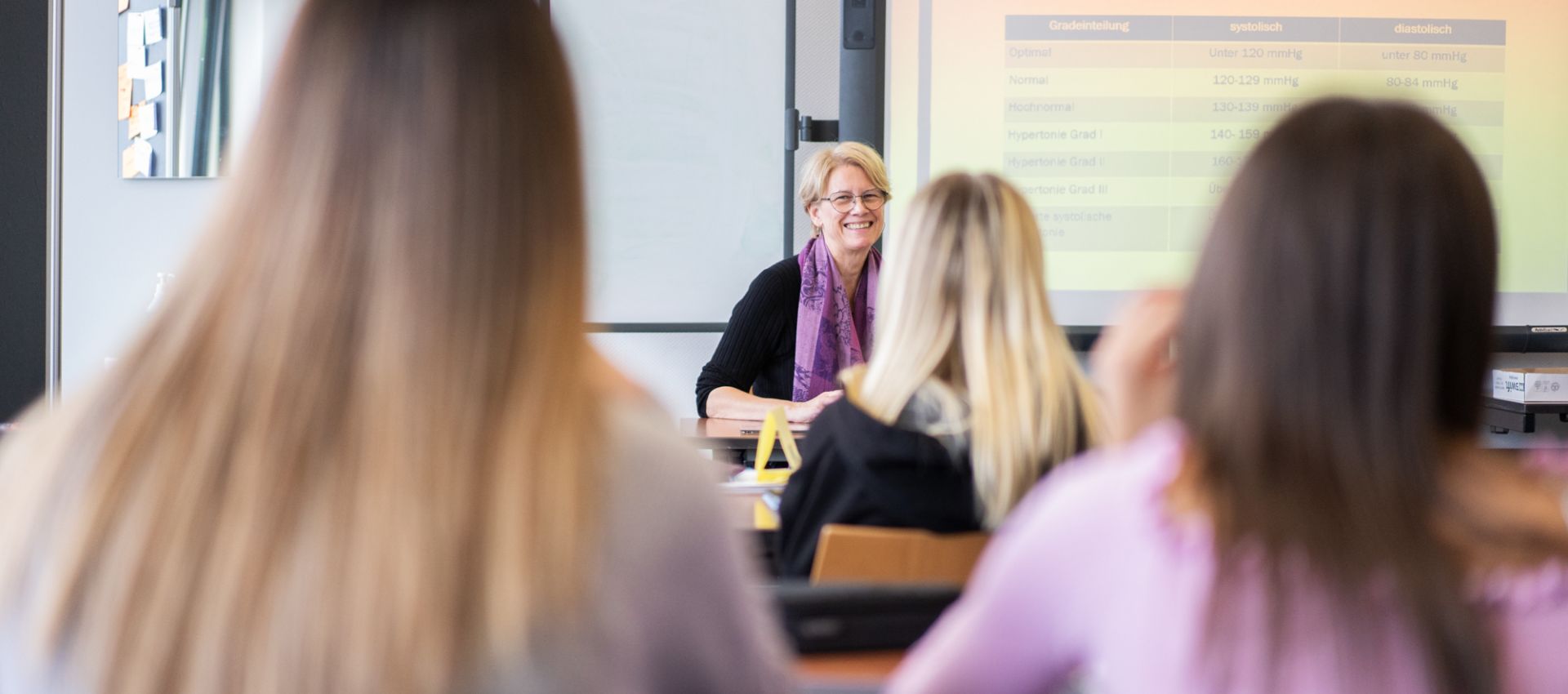 Im Klassenraum der Pflegeschule sitzt eine Lehrkraft vor einer Projektionsfläche mit Präsentationsfolie. Mehrere Auszubildende blicken zur Tafel. Die Stimmung wirkt konzentriert und freundlich.