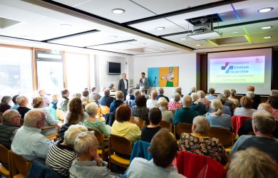 Zahlreiche Besucherinnen und Besucher sitzen in einem Veranstaltungsraum. Sie sitzen in Stuhlreihen nebeneinander und sind von hinten zu sehen. Sie verfolgen einen Vortrag mit Präsentation.