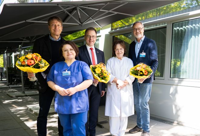 Fünf Personen stehen auf der Terrasse der Palliativstation. Drei halten Blumensträuße in den Händen, sie tragen Alltags- und Businesskleidung. Zwei Mitarbeiterinnen tragen Pflegekleidung. Im Hintergrund sind Sonnenschirme zu sehen.