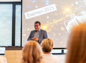 Vorn am Rednerpult spricht Franz Hafner, kaufmännischer Geschäftsführer EVKLN, vor einer Leinwand mit dem Schriftzug ‚Examen‘ und Sektgläser-Projektion; im Vordergrund sind einige Zuhörer von hinten zu sehen.