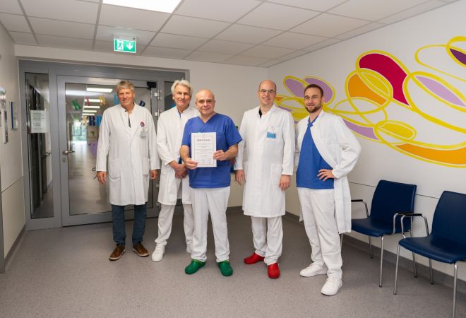 Gruppenbild auf dem Stationsflur. 5 Ärzte stehen nebeneinander, einer von ihnen hält ein Zertifikat in die Kamera. Alle lächeln. Im Hintergrund ist eine gelb-rote Wandmalerei zu sehen.