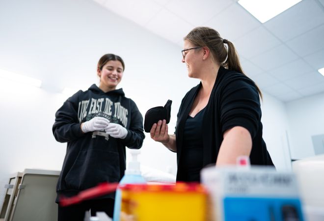 Eine Ausbilderin zeigt einer Auszubildenden etwas auf einem Tablet. Beide lächeln. Die Auszubildende trägt Einweghandschuhe, im Vordergrund sind Pflegeutensilien unscharf zu sehen. Die Szene zeigt eine positive Lernatmosphäre im Praxisunterricht.