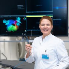 Eine Ärztin im weißen Kittel steht in einem Behandlungsraum und hält einen Katheter in der Hand. Sie blickt lächelnd in die Kamera. Hinter ihr ist ein Monitor mit einer farbigen medizinischen Darstellung zu sehen.