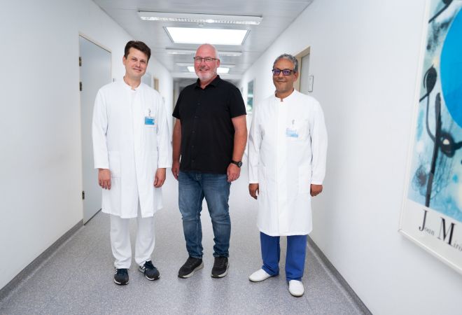 Drei Männer stehen in einem hellen Krankenhausflur. In der Mitte steht ein Mann in Alltagskleidung. Links und rechts steht jeweils ein Arzt. Die Ärzte tragen einen weißen Kittel. Alle drei lächeln.