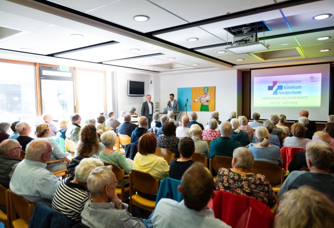 Zahlreiche Besucherinnen und Besucher sitzen in einem Veranstaltungsraum. Sie sitzen in Stuhlreihen nebeneinander und sind von hinten zu sehen. Sie verfolgen einen Vortrag mit Präsentation.