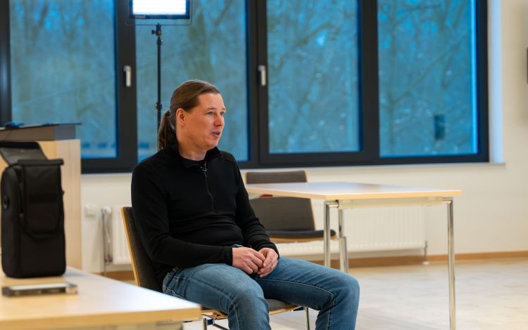 Ein Mann sitzt auf einen Stuhl in einem großen, hellen freundlichen Raum. Im Hintergrund ist eine große Fensterfront zu sehen. Hinter ihm steht ein Scheinwerfer. Er ist im Gespräch.
