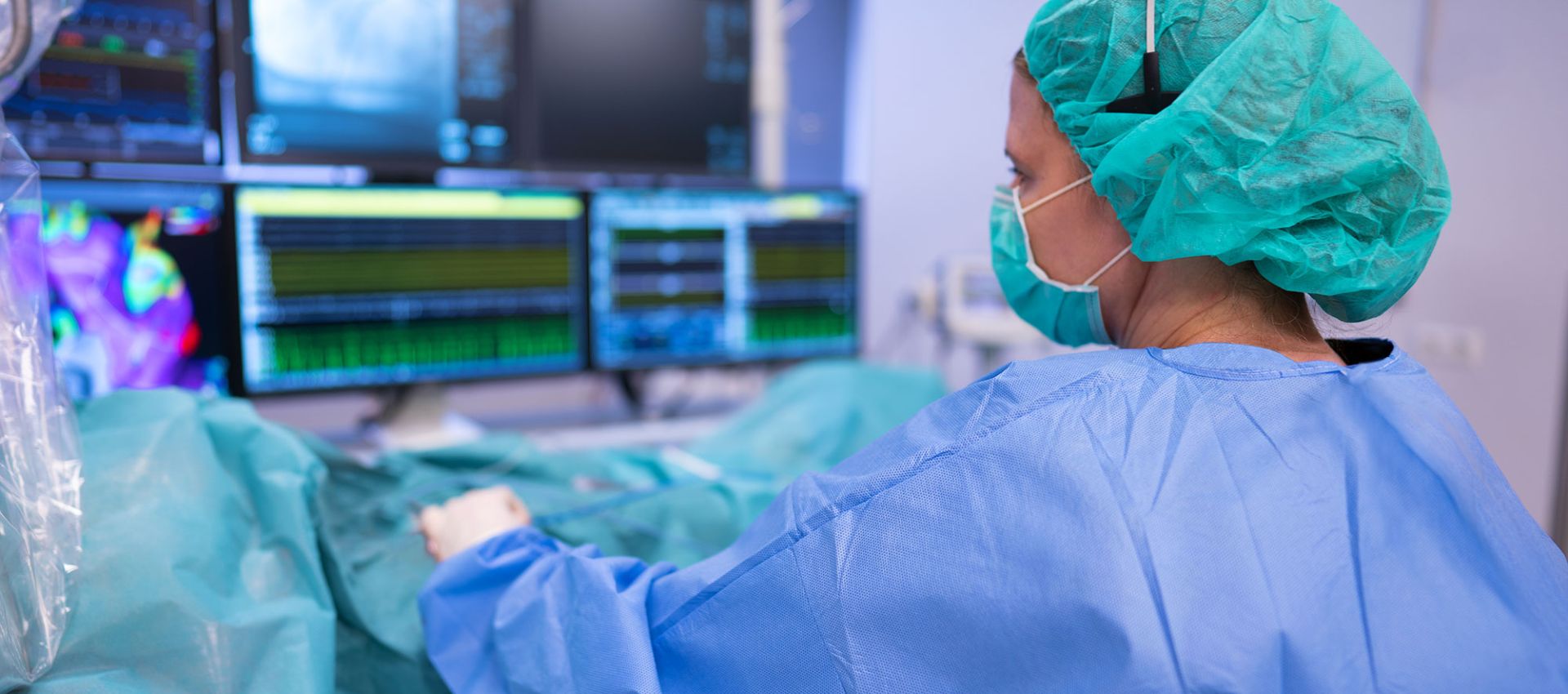 Dr. Ute Ruprecht führt einen kathetergestützten Eingriff am Herzen durch. Sie trägt sterile OP-Kleidung und schaut konzentriert auf mehrere Monitore, die gegenüber stehen.
