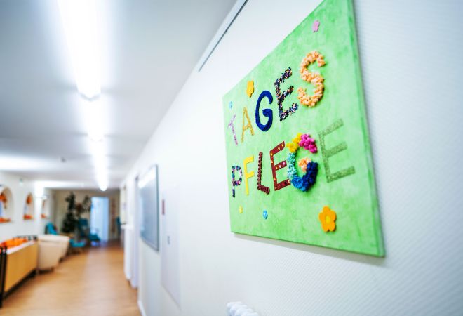 An einer hellen Wand im Eingangsflur der Tagespflege hängt auf Augenhöhe ein handgemachtes grünes Schild mit der Aufschrift „Tagespflege“. Die bunten Buchstaben bestehen aus verschieden gemusterten Stoffen und wirken einladend und liebevoll gestaltet.