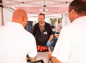 Marc-André Dost, pflegerischer Leiter der Zentralen Notaufnahme, steht hinter einem Tisch unter dem Pavillon und bereitet einen Burger zu. Vor ihm stehen zwei Männer aus dem Rettungsdienst, die sich bei der Grillaktion einen Imbiss holen.