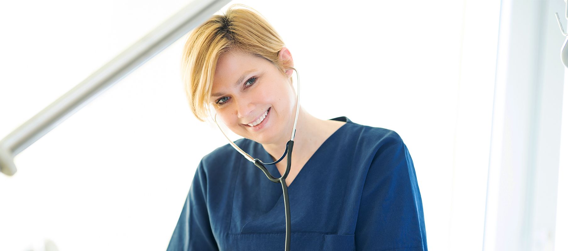 Eine weibliche Pflegekraft steht am Patientenbett und nutzt ein Stethoskop, um die Vitalzeichen des Patienten zu überprüfen.