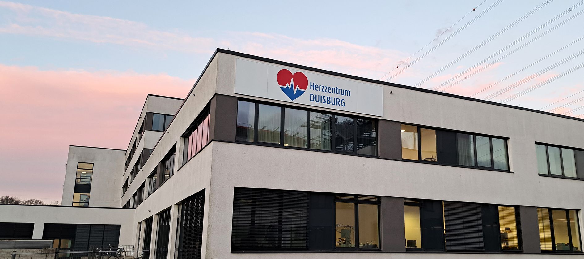 Außenansicht des Herzzentrums Duisburg am Standort in Duisburg Fahrn. An der Fassade ist das Logo des Herzzentrums angebracht. Der Himmel im Hintergrund ist rosa vom Sonnenaufgang.