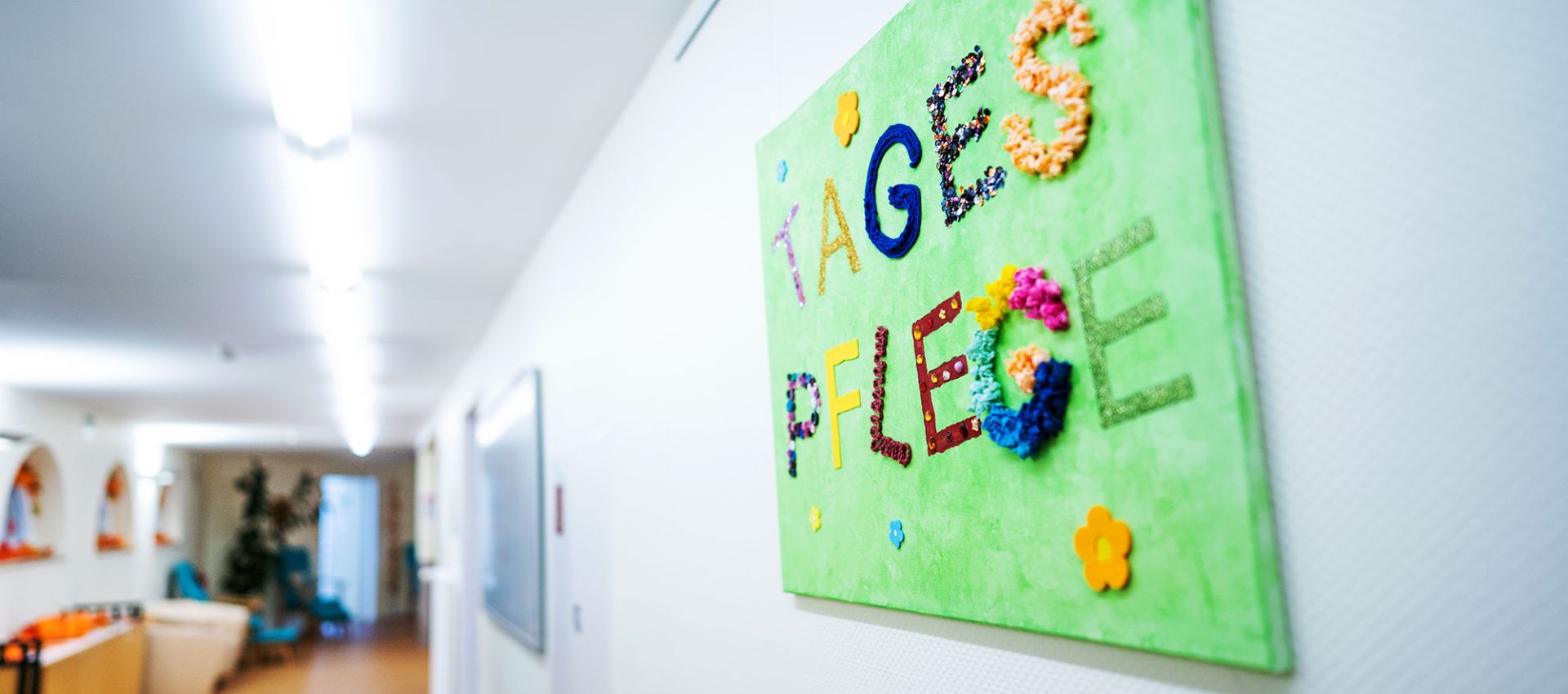 An einer hellen Wand im Eingangsflur der Tagespflege hängt auf Augenhöhe ein handgemachtes grünes Schild mit der Aufschrift „Tagespflege“. Die bunten Buchstaben bestehen aus verschieden gemusterten Stoffen und wirken einladend und liebevoll gestaltet.