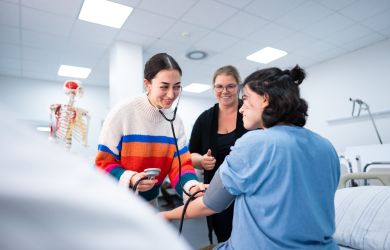 Eine Auszubildende misst im Praxisunterricht der Pflegeschule den Blutdruck bei einer Mitschülerin. Alle Beteiligten lächeln, im Hintergrund steht die Ausbilderin unterstützend dabei.
