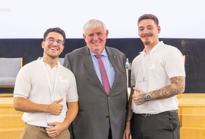 Gruppenfoto beim Fachtag „Zukunftsbündnis Pflege“: In der Mitte NRW-Gesundheitsminister Karl-Josef Laumann, flankiert von zwei jungen Pflegeauszubildenden in weißem Poloshirt. Alle drei lächeln, im Hintergrund eine Bühne mit Leinwand.