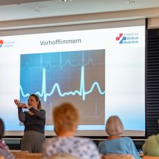 Dr. med. Ute Ruprecht steht vor einem Publikum und hält einen Vortrag über Vorhofflimmern. Hinter ihr ist eine große Präsentation mit einer Herzkurve zu sehen.