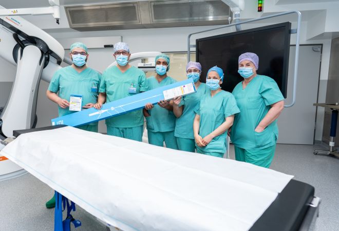 Mehrere Mitarbeitende eines OP-Teams stehen in grüner Schutzkleidung und mit Mundschutz in einem Eingriffsraum neben einem Behandlungstisch. Zwei Personen halten die Verpackung eines medizinisches Produktes in den Händen.