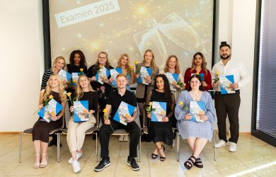 Gruppenfoto von 13 Absolvent*innen einer Pflegeausbildung vor einer Leinwand mit der Aufschrift „Examen 2025“. Alle halten stolz ihre Urkunde und eine gelbe Rose in den Händen und lächeln in die Kamera.