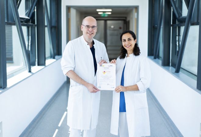 Ein Arzt und eine Ärztin präsentieren in einem Verbindungsgang des Krankenhauses das Zertifikat zur Auszeichnung des Evangelischen Klinikums Niederrhein als Cardiac Arrest Center.