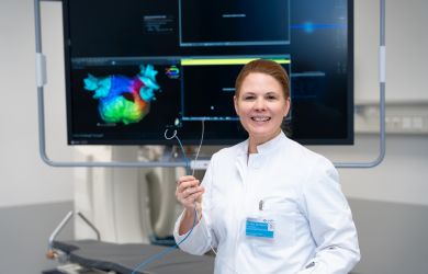 Eine Ärztin im weißen Kittel steht in einem Behandlungsraum und hält einen Katheter in der Hand. Sie blickt lächelnd in die Kamera. Hinter ihr ist ein Monitor mit einer farbigen medizinischen Darstellung zu sehen.
