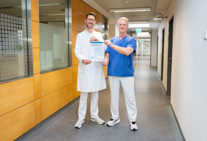 Dr. Philipp Weber (links) und Kersten Brenner (rechts) stehen lächelnd in einem Krankenhausflur und halten gemeinsam das Zertifikat des Regionalen Traumazentrums in die Kamera. Weber trägt einen weißen Arztkittel, Brenner blaue OP-Kleidung.