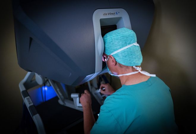 Prof. Dr. Börgermann in OP-Kleidung sitzt an der Steuerkonsole des Da-Vinci-Operationssystems und blickt durch das Okular. Aufnahme von hinten während eines Eingriffs.