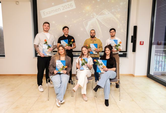 7 Absolventinnen und Absolventen der Pflegeschule, eine Reihe sitzend, dahinter weitere stehend; alle halten Zeugnis-Mappe und Rose, im Hintergrund Leinwand mit ‚Examen 2026‘.