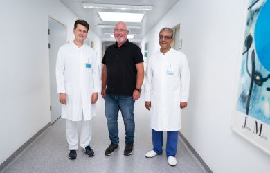 Drei Männer stehen in einem hellen Krankenhausflur. In der Mitte steht ein Mann in Alltagskleidung. Links und rechts steht jeweils ein Arzt. Die Ärzte tragen einen weißen Kittel. Alle drei lächeln.