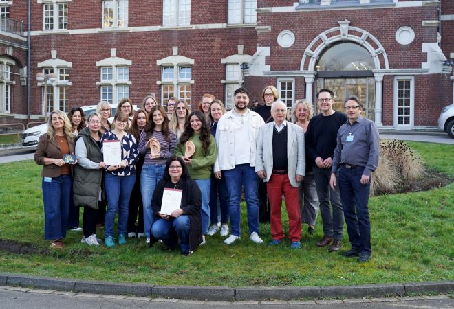 Eine Gruppe von rund zwanzig Teilnehmenden der Akupunktur-Weiterbildung steht gemeinsam auf einer Wiese vor dem historischen Gebäude des Johanniter-Krankenhauses Oberhausen. Einige halten Zertifikate oder Anschauungsmodelle eines Ohrs hoch und lächeln.