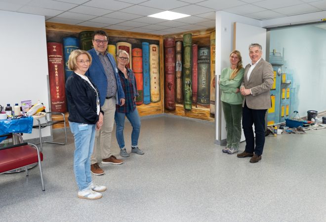 Mehrere Personen stehen im Klinikflur vor einer fertig gestalteten Wand mit großformatigen, bunten Bücher-Motiven. Zwei der beteiligten Mitarbeiterinnen stehen daneben, Malutensilien sind im Hintergrund zu sehen.