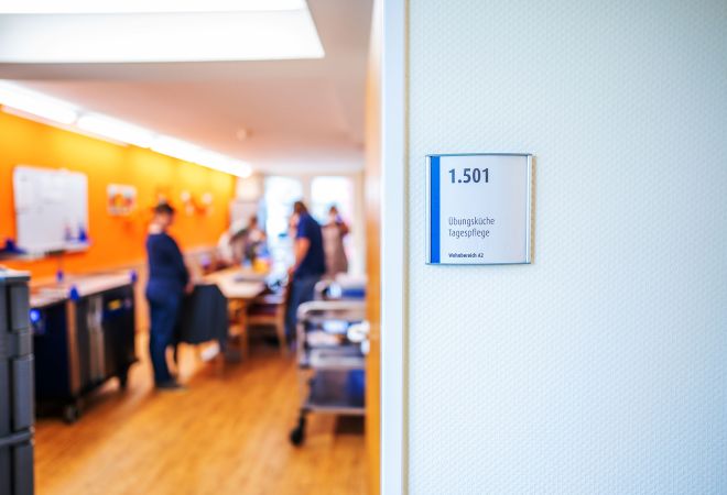 Das Bild zeigt einen Blick durch eine geöffnete Tür in die Therapieküche der Tagespflege. Am Türrahmen hängt ein Schild mit der Raumnummer 1.501 und der Aufschrift „Übungsküche Tagespflege“. Im Raum sind mehrere Personen zu sehen. Die Wände sind orange.
