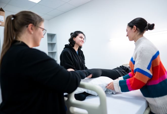 In einem Übungsraum der Pflegeschule liegt eine Auszubildende auf einem Pflegebett. Zwei weitere Auszubildende stehen daneben, am Fußende beobachtet eine Ausbilderin die Szene. Die Gruppe übt eine Pflegesituation in entspannter Lernatmosphäre.