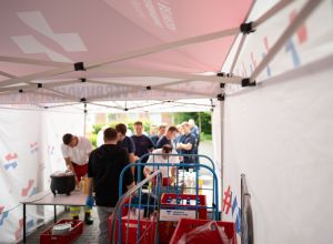 Blick aus dem Pavillon bei der Grillaktion am Evangelischen Krankenhaus Dinslaken: Im Vordergrund stehen Küchenwagen mit Kisten und eine Tischreihe. Davor holen sich mehrere Rettungskräfte in einer Schlange etwas zu essen ab.