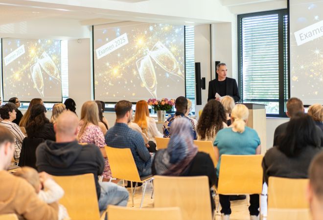 Blick in den Saal der Examensfeier der Pflegeschule; von hinten auf die Sitzreihen mit Auszubildenden und Angehörigen, vorne spricht Torsten Edelkraut am Rednerpult, dahinter Leinwände mit dem Schriftzug „Examen“ und projizierten anstoßenden Sektgläsern.