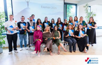 18 Absolventinnen und Absolventen der Pflegeschule, eine Reihe sitzend, dahinter weitere stehend; alle halten Zeugnis-Mappe und Rose, im Hintergrund Leinwand mit ‚Examen 2026‘.