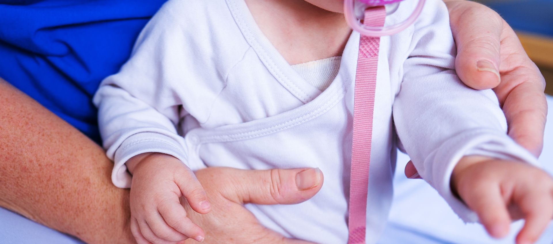 Ein Baby mit pinkfarbenem Schnuller sitzt auf dem Boden. Die Physiotherapeutin umfasst das Baby sanft von hinten. Das Gesicht des Babys ist nicht vollständig sichtbar.