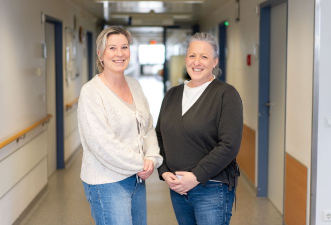 Die Pain Nurses Corinna Noy und Annika Trapphoff stehen nebeneinander in einem Krankenhausflur und lächeln in die Kamera.